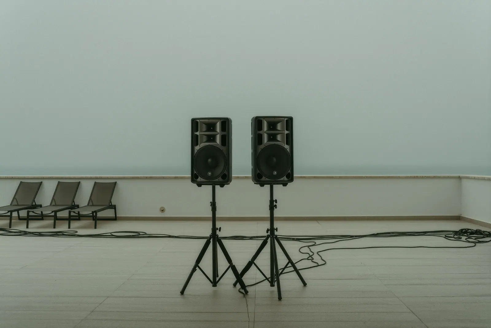 A pair of PA speakers on tripod stands facing an empty villa terrace under flat grey light, cables taped down across the tiles and folded loungers behind.
