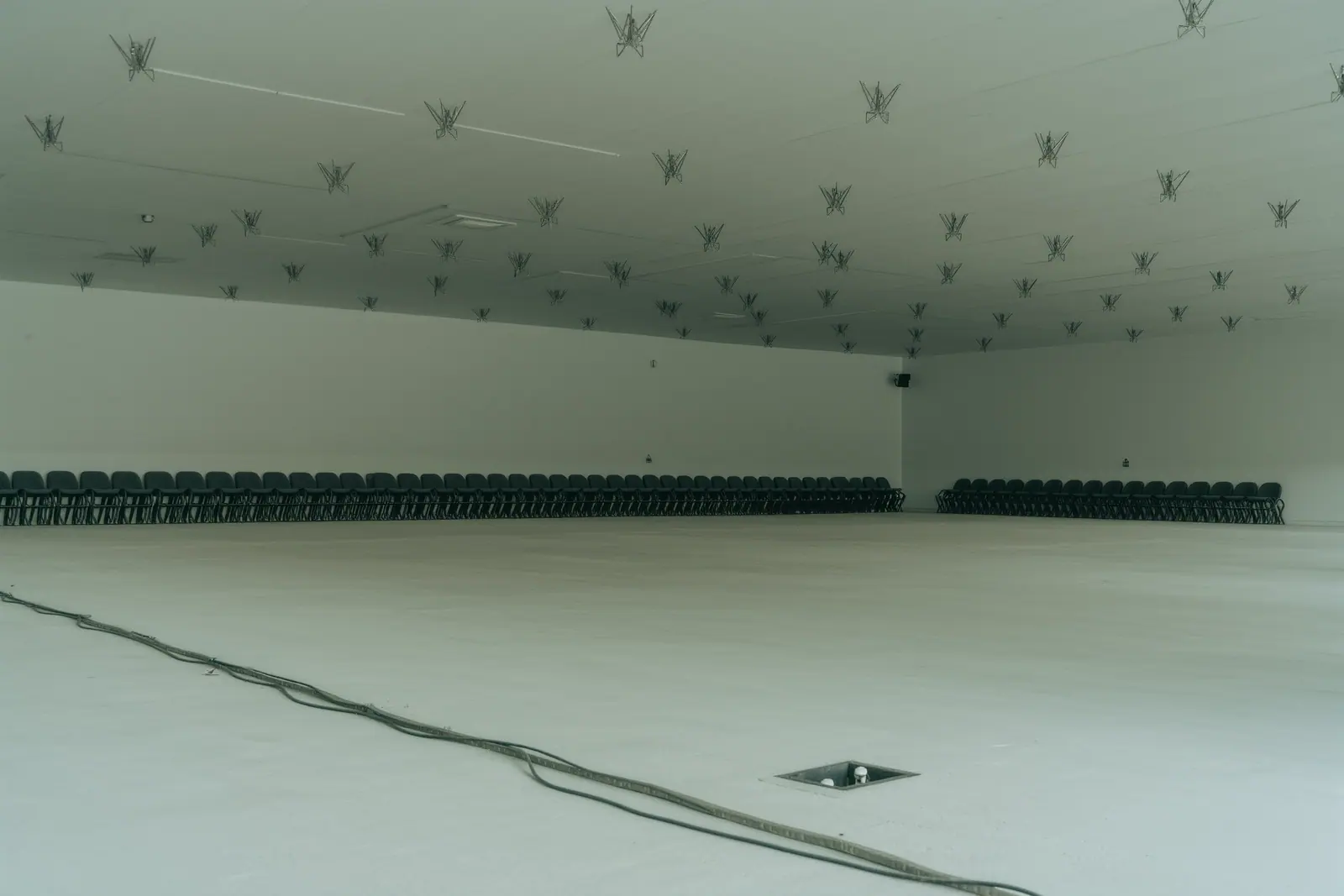 An empty whitewashed event space with bare steel rigging points in the ceiling, stacked seating against a wall and a taped-down cable run across the floor.