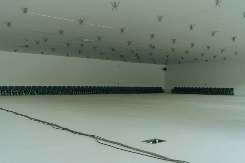 An empty whitewashed event space with bare rigging points in the ceiling and stacked seating against a wall.