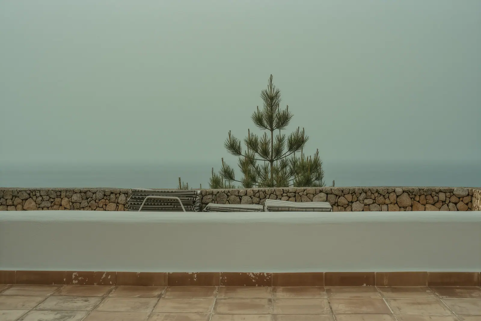The empty terrace of a whitewashed Ibizan villa with sun loungers folded and stacked against a low wall, chipped terracotta tiles underfoot and sabina pine beyond.