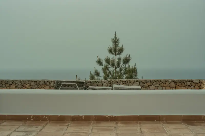 The empty terrace of a whitewashed Ibizan villa in low light, folded sun loungers, a dry stone wall and pine beyond.