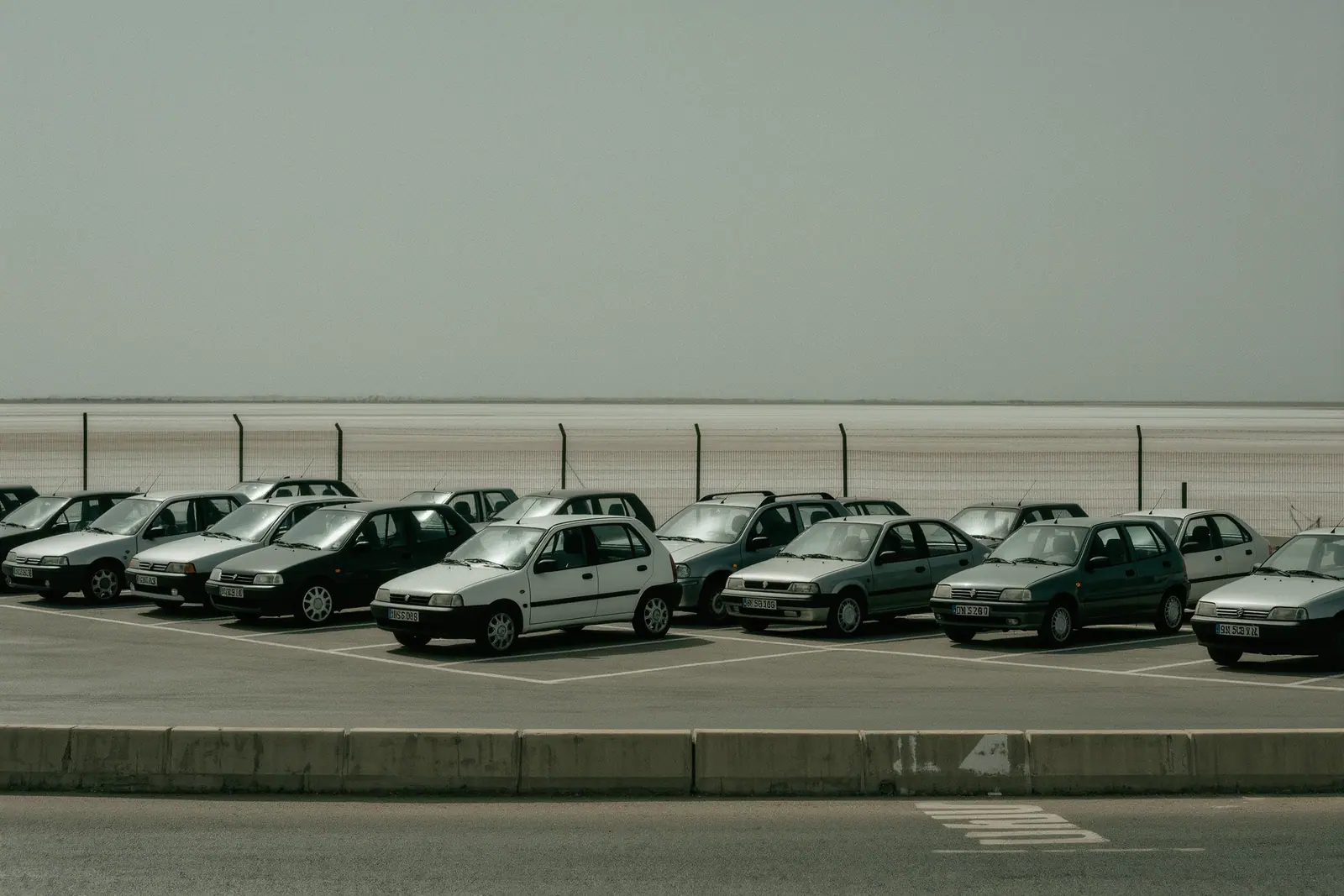 An open rental car lot near Ibiza airport with small unbranded cars in numbered bays, a painted bay number on the tarmac and a wire fence over flat salt-flat land beyond.