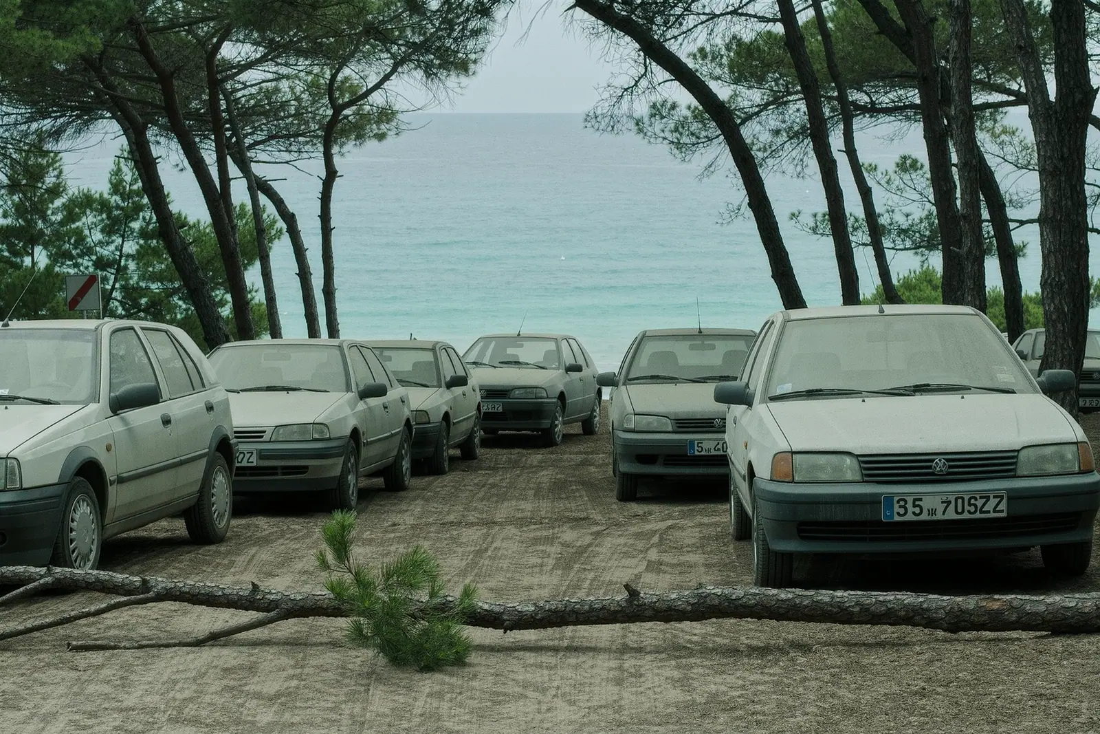 A dirt overflow car park above a busy Ibizan cala with dusty cars parked nose-to-tail under thin pines and dry rutted earth in the foreground.