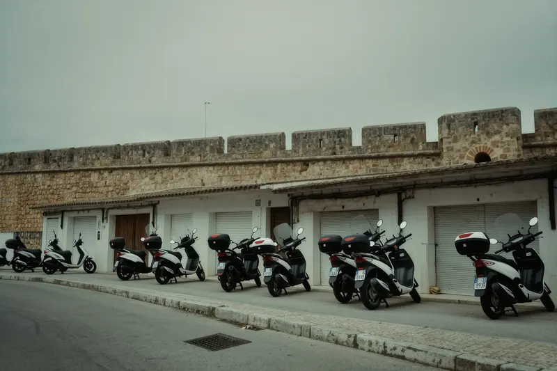 Scooters parked nose-in along a whitewashed one-way street, front wheels turned to the kerb, wall shadow across the road.