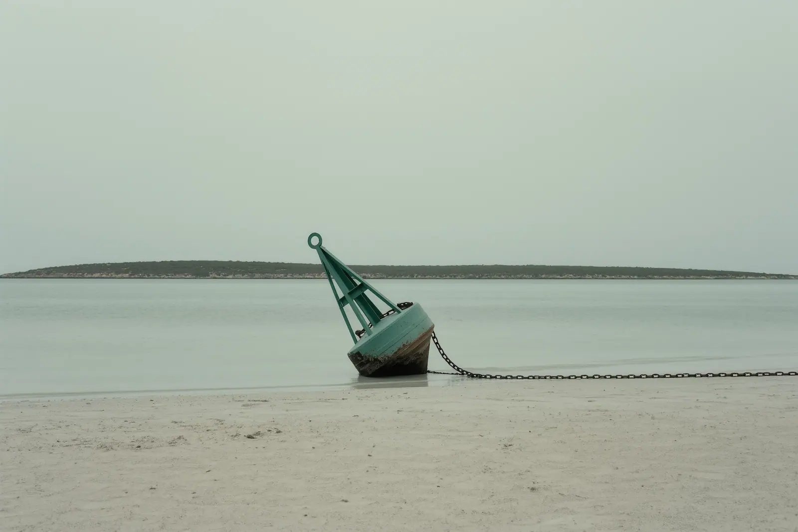 The flat turquoise shallows of the Es Freus channel between Ibiza and Formentera with a weathered channel marker buoy leaning in the foreground and no boats in sight.