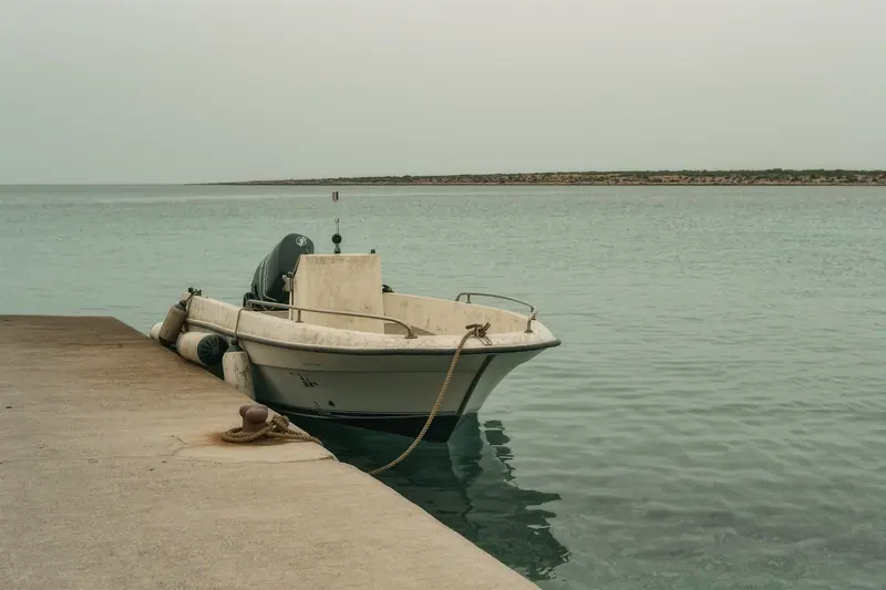 A small open speedboat tied alongside a concrete jetty, worn fenders compressed between the hull and the wall, salt-crusted rope running to a cleat.