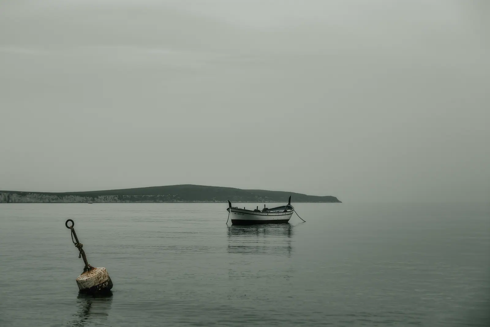 A small day boat riding on a mooring buoy off the west coast of Ibiza under flat grey cloud, the weathered buoy and its slack pick-up line close in the water.
