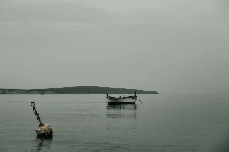 A small day boat lying to a mooring buoy off a low Ibizan shoreline under flat grey cloud.